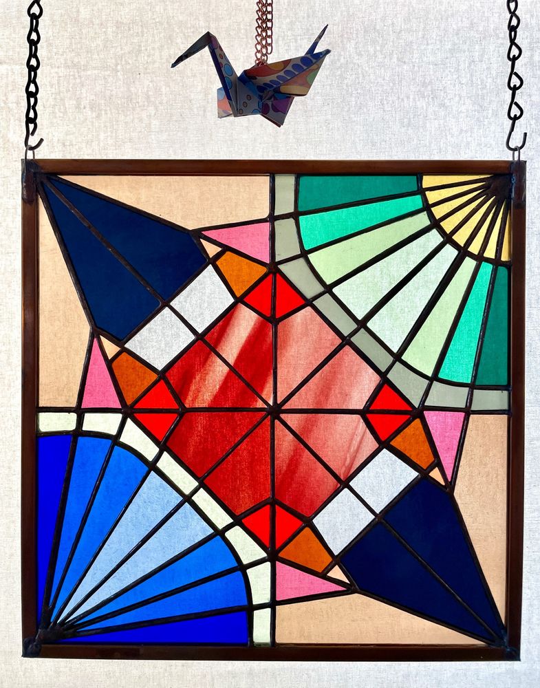 a square stained glass panel with very rich, vibrant jewel toned geometric pieces. It’s divided into four quadrants. The upper right and lower left have radiating lines in them, representing the crane’s head and tail sections. The upper left and lower right have larger dark purple triangles that form the crane’s wings 