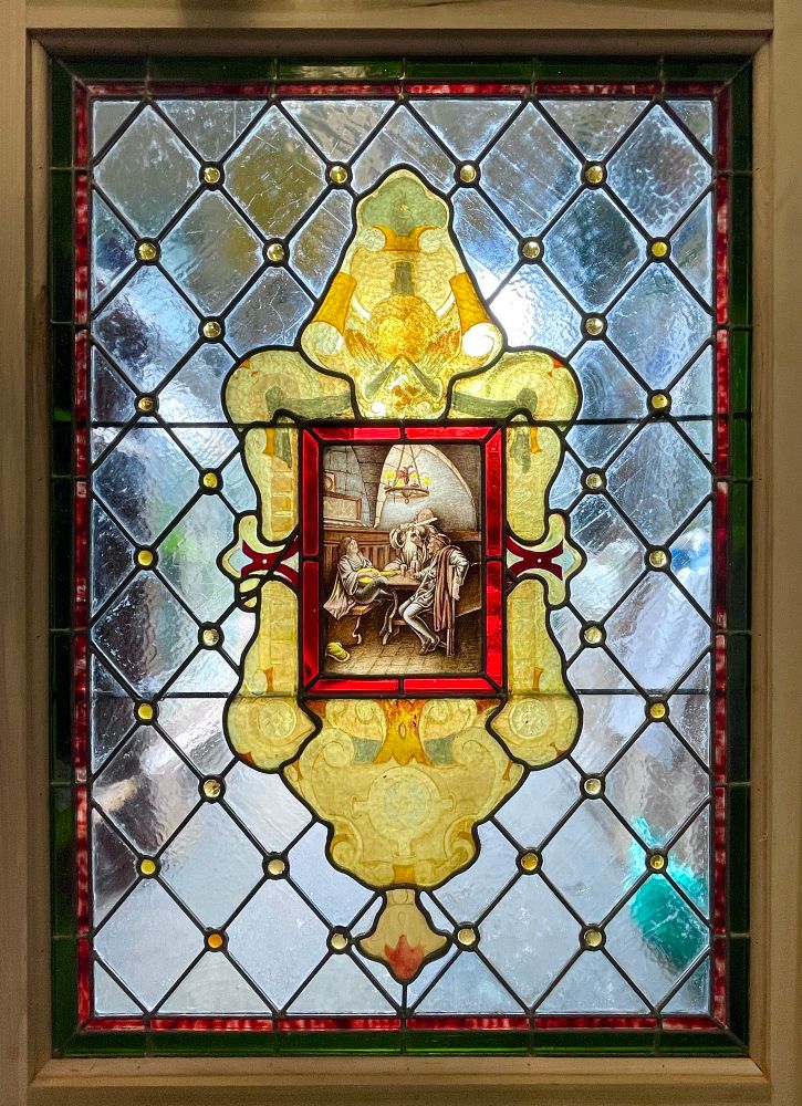 an early 19th-century stained glass window with a pale blue diamond pane background. small round amber gems dot each intersection in the background. there's a double border, a thicker dark green on the outside and a thin red pencil border on the inside. the center is a rectangular painted panel depicting 3 men in a tavern playing music and drinking. it is surrounded by a red border and the whole is surrounded by an ornate border of painted amber scrollwork 