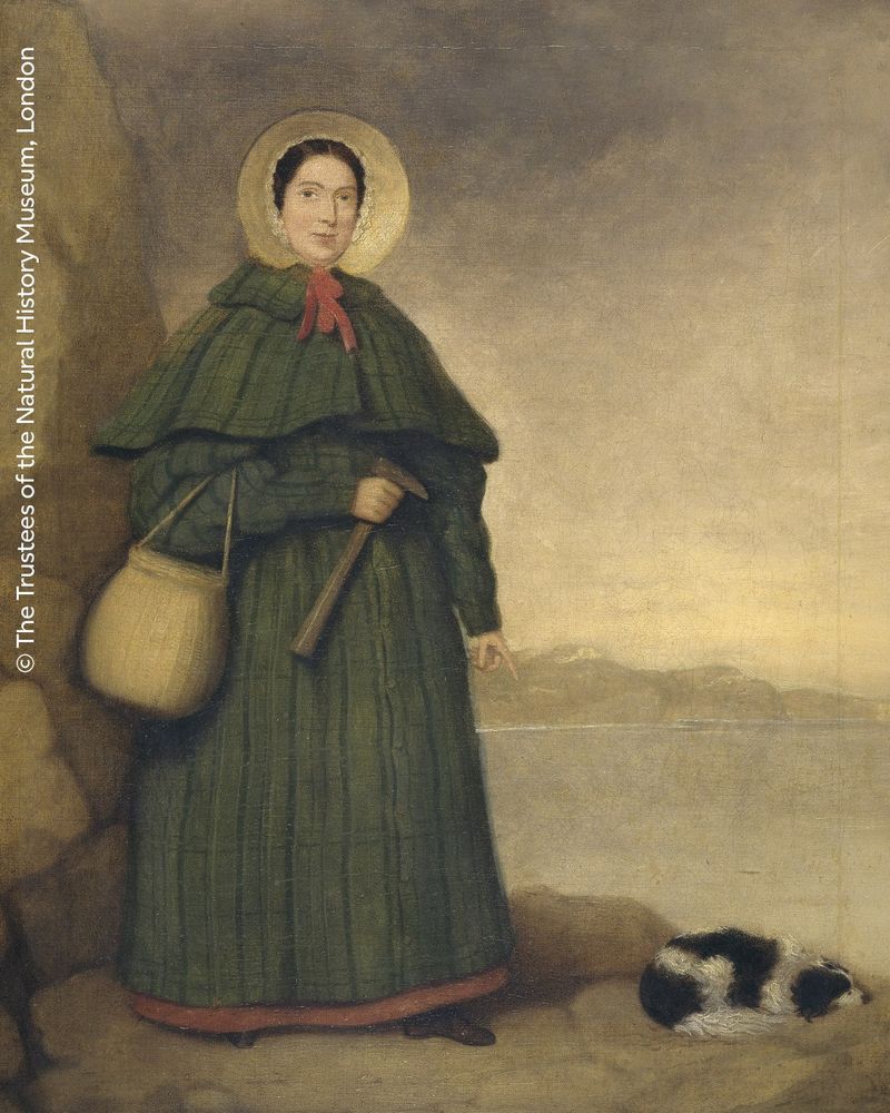 A painting of a portrait of the palaeontologist Mary Anning. She is wearing a long, tiered dark dress, a bonnet and a red scarf. She’s holding a wicker basket around her elbow and a hold a hammer in her hand. Her other hand points to a small dog on the ground. 
