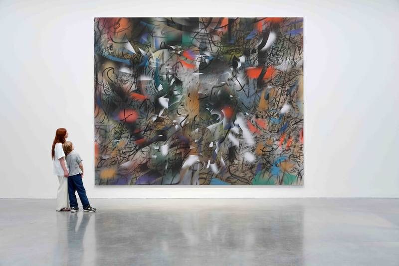 A large brightly coloured painting with two people standing to the left looking up at the work. The wall behind is white.