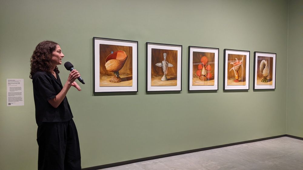 Young woman wearing black, standing with a microphone to the left of five wall mounted framed paintings.