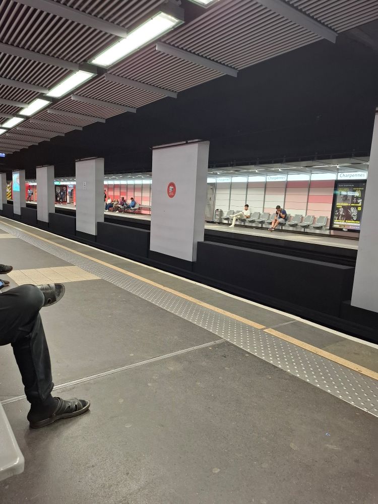 Station de métro, on voit des jambes dépasser à gauche, sur la droite il y a les voies et on aperçoit le quai d'en face avec le mur rose et blanc (le rose est la couleur de la ligne) il y a des gens assis sur les assises 