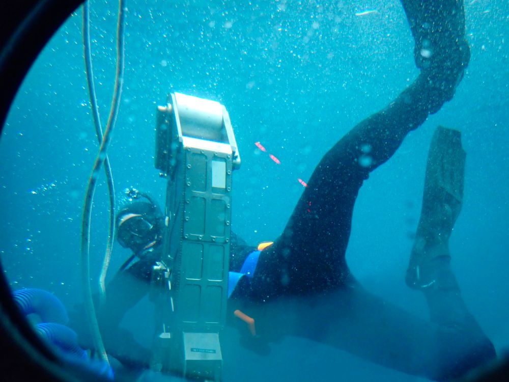Swimmers in a wetsuit attaching a line to the front of the HOV Alvin. One of Alvin’s manipulator arms is visible. Water near the surface was a beautiful deep turquoise. Photo courtesy of Amanda Demopoulos, USGS; NOAA Ocean Exploration, ONR, NOPP, BOEM, NOAA IOCM, USGS; ©Woods Hole Oceanographic Institution