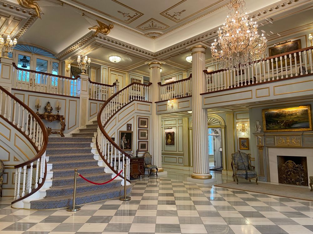 The grand foyer with a symmetric staircase, a checkerboard white and gray marble floor, a wrap around balcony overlooking the main foyer, and several chandeliers and fireplaces.