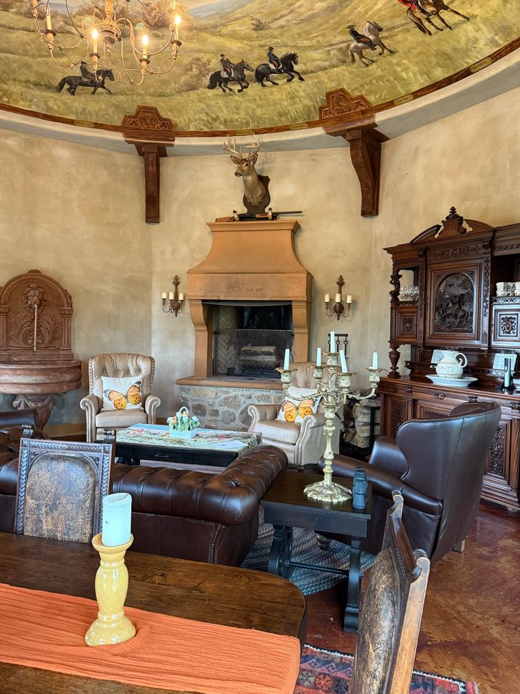 The British Folly on the back property serves as a hunting room with brown leather furniture, a rustic fireplace, and a fox hunting theme mural painted on the dome ceiling. The tones are warm- in contrast to the to the cool tones of the main house.
