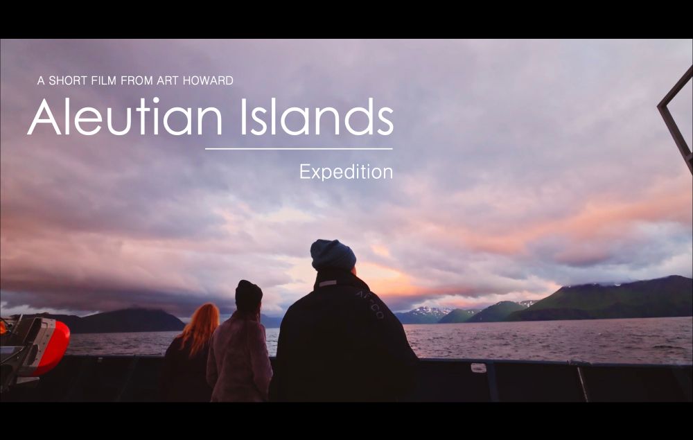 Film cover showing three ocean expedition members standing on this stern of a research vessel viewing the Aleutian Islands. The clouds are reflecting the low sun’s pastel shades of blue, pink, and purple