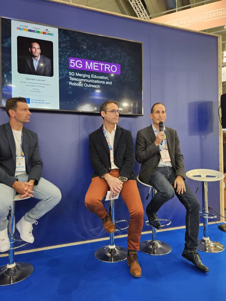 Three men seated on high stools at a professional conference, in front of a blue backdrop. The man on the right, Sylvain Leroux, is the representative of the 5G Metro project. He is holding a microphone and speaking. Behind them, a screen displays the title “5G METRO” and the name “Sylvain Leroux” along with a portrait photo. The scene is part of a presentation of the 5G Metro project at the European Commission booth during the Smart City Expo World Congress.