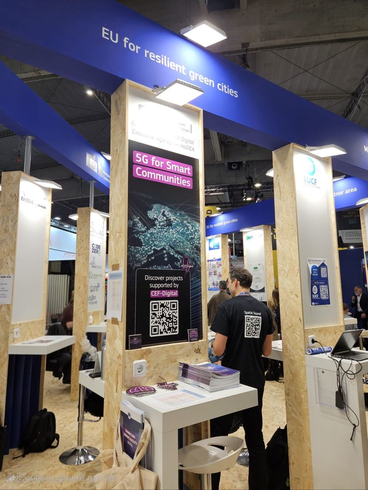 Photo of an exhibition booth themed “EU for Green and Resilient Cities.” The booth highlights the “5G for Smart Communities” theme supported by CEF Digital, featuring a large poster with a QR code and brochures on the counter. Several other booths and visitors are visible in the background, under a wooden structure and hanging blue signs.