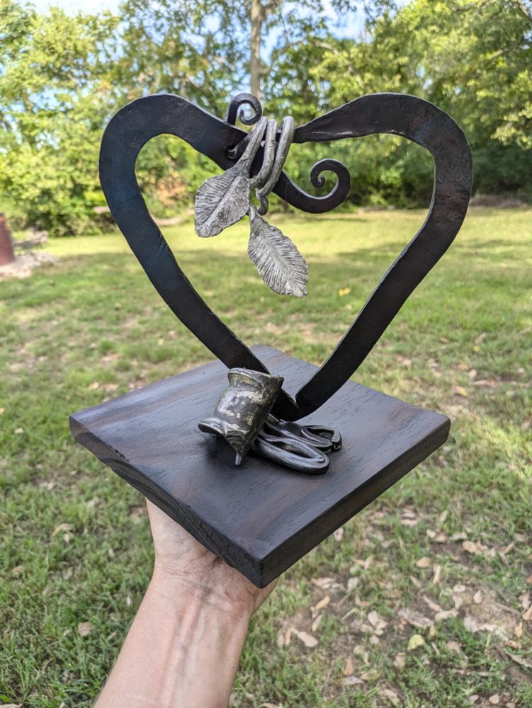 Sculpture of a metal heart with long-stemmed aluminum leaves wrapped around the top and some curled steel at the base. Mounted on a wooden plank. Background is a grassy area with trees.
