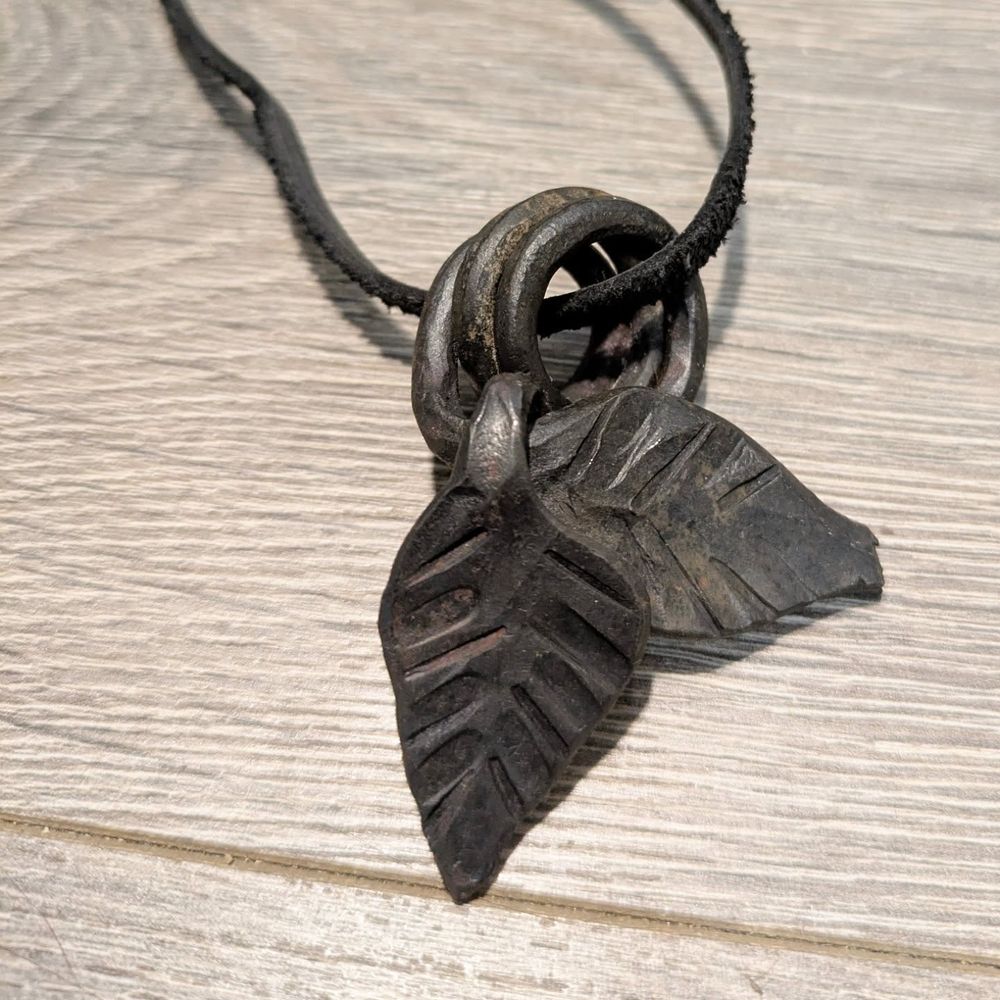 Two leaves with coiled stems made from steel, with a leather lace going through the coils. Wood textured background.