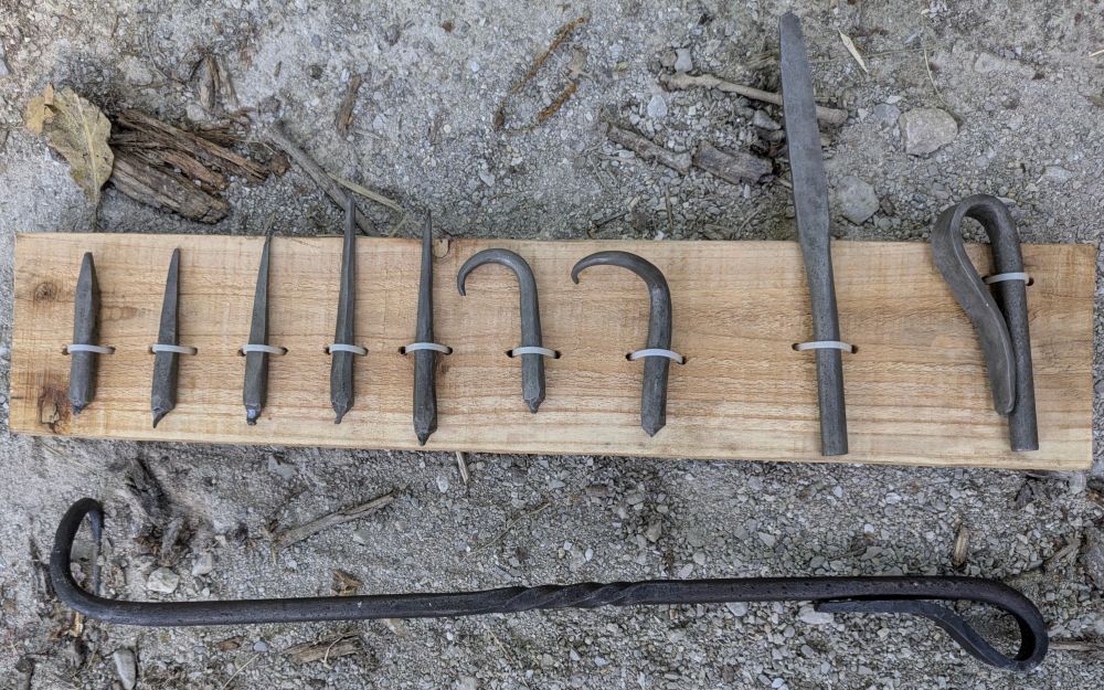 Example showing progressive steps for making a pigtail bbq flipper out of steel. Pieces are attached to a board and a finished flipper is laying along the base. Gravel and twigs and leaves in the background.