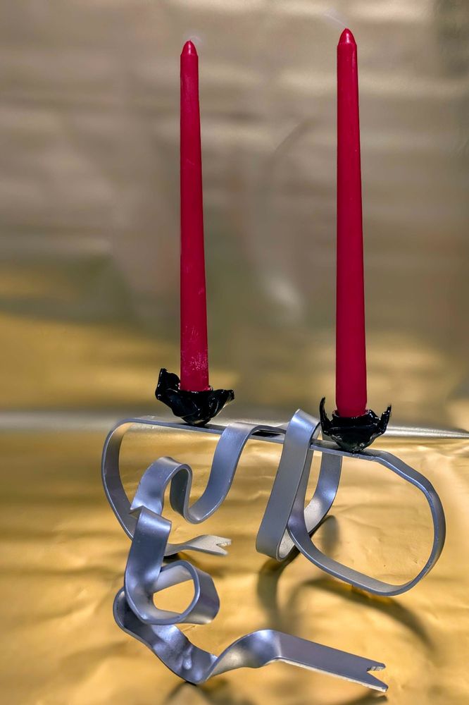 Two red taper candles sit in a metal candle holder base which looks like ribbon. The cups which the candles are sitting in are black. The background is gold.
