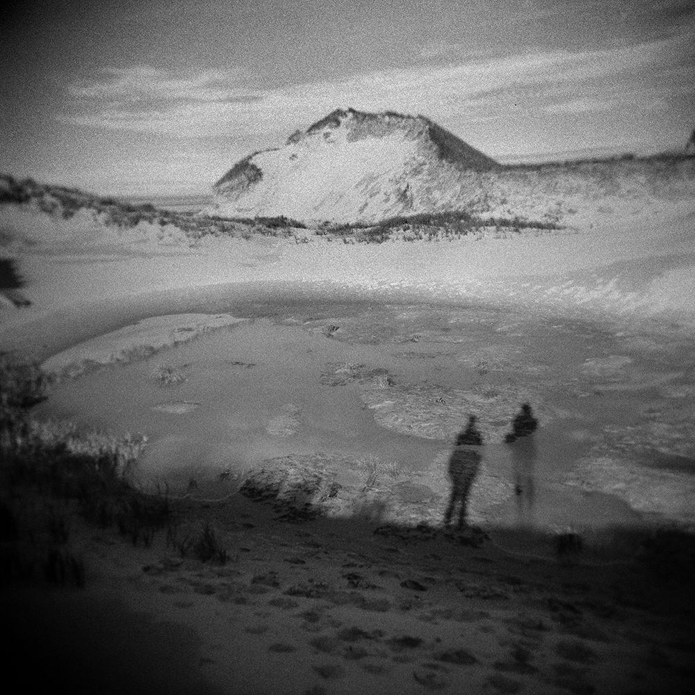 Black and white square photo. The shadows of two figures stand on the edge of coastal sand dunes. There is an empty, bowl-shaped area in front of them and a large dune in the distance.