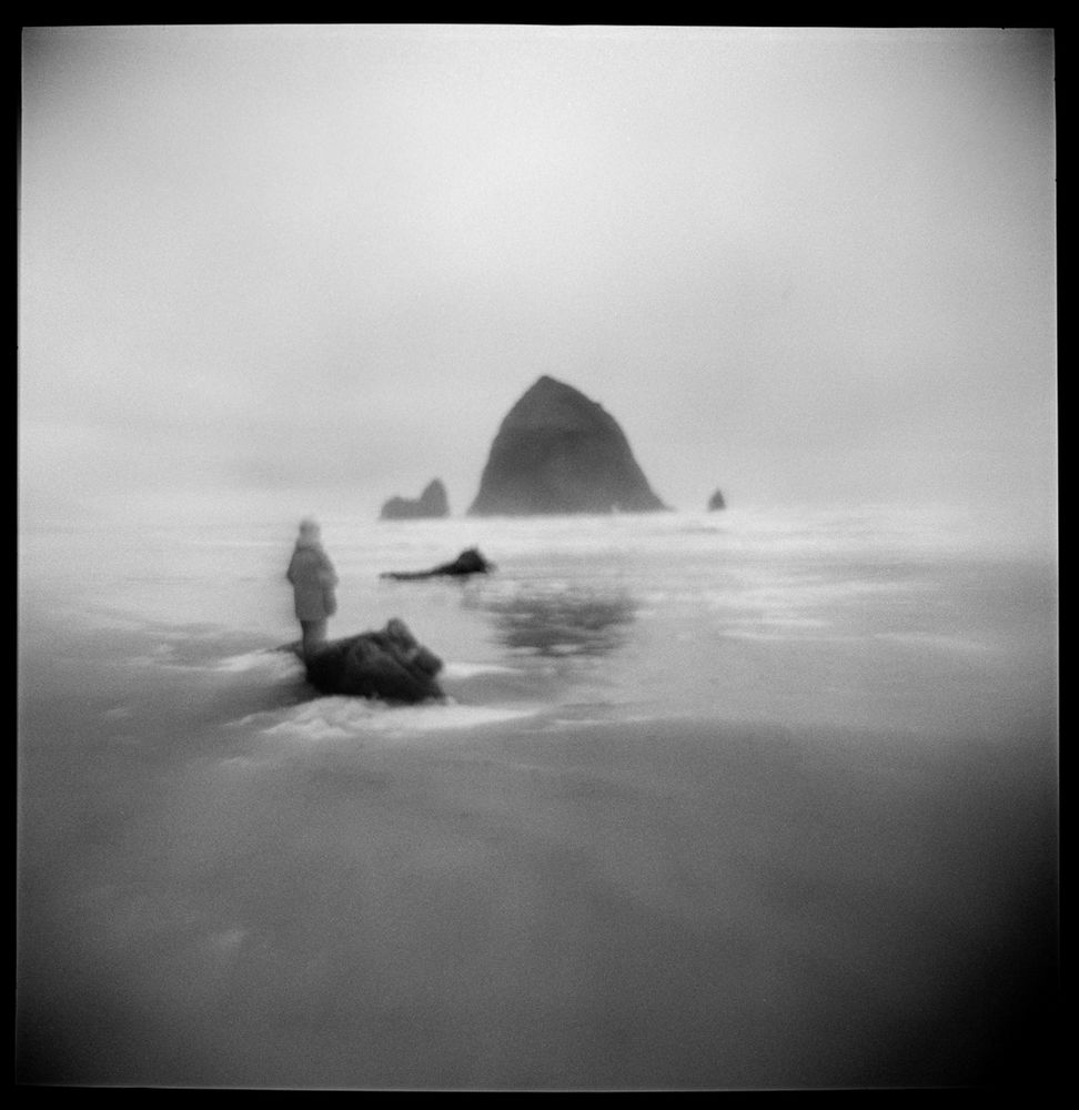 A black and white square image mad at Cannon Beach on the Oregon coast. A sole figure stands on the shore looking out at a somewhat triangular rock formation (Haystack Rock) just off of the coast. The image has a soft, overall blur to it from using a plastic lens that had been inverted (or "flipped").