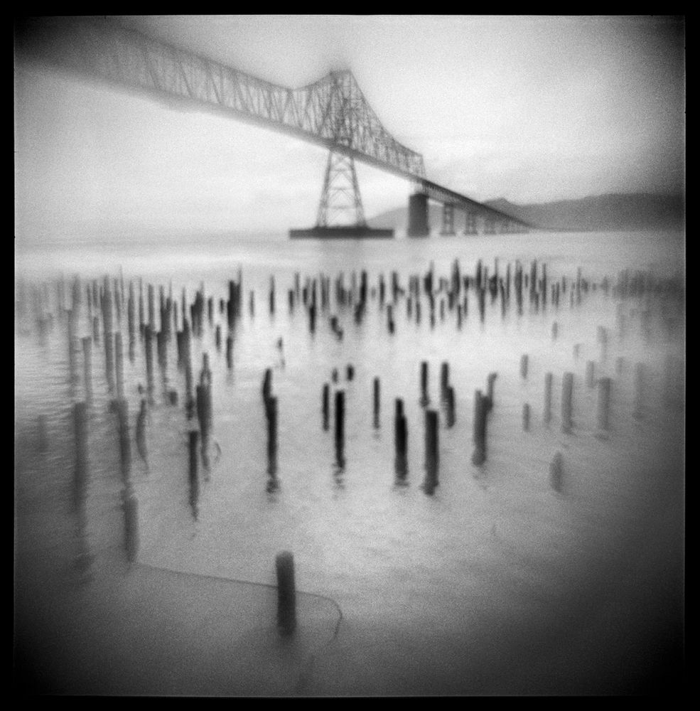 Black and white, square image. A bridge spans the Columbia River in Astoria, Oregon on the Pacific coast. Pilings from an old pier dominate the foreground. The image has an overall blur to it; the result of using a cheap plastic lens that was inverted before placing it in the camera.