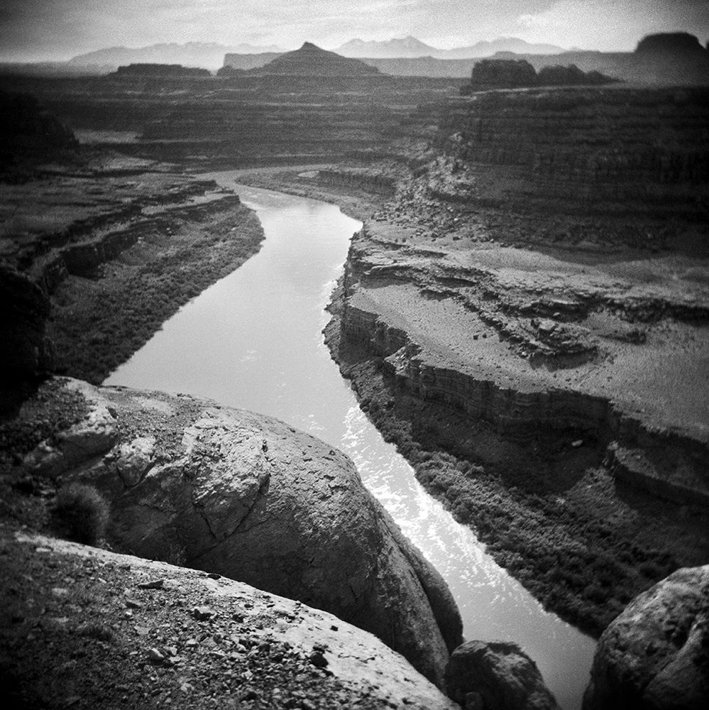 A black and white square image made while standing on a cliff's edge looking hundreds of feet down at the Colorado River as it winds its way through the distant canyons and buttes of Canyonlands National Park in southern Utah.