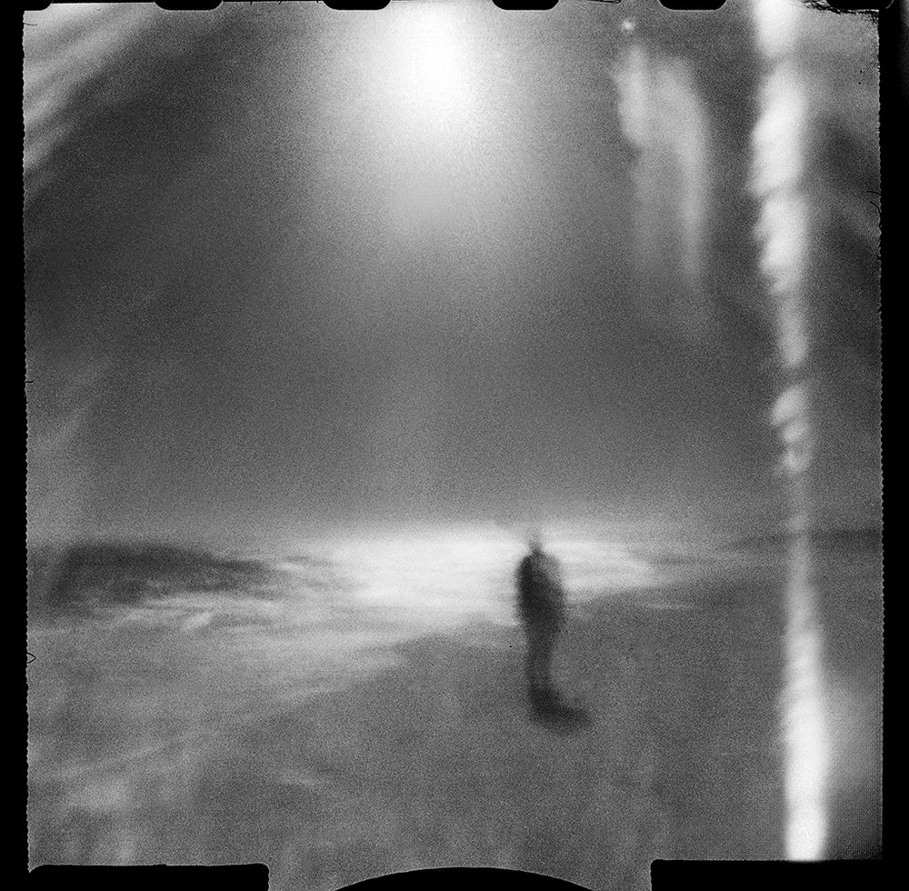 Black and white square photo made with a small 35mm pinhole camera that takes 24x24 square images. A person stands alone along the ocean shore. The camera is pointed at the sun which is causing large amounts of flare to dominate the scene. 