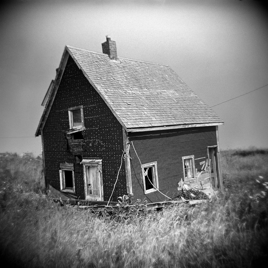 Black and white square image made with a Holga plastic camera. A small abandoned house sits somewhat tilted in the dune grass near the ocean shore.
