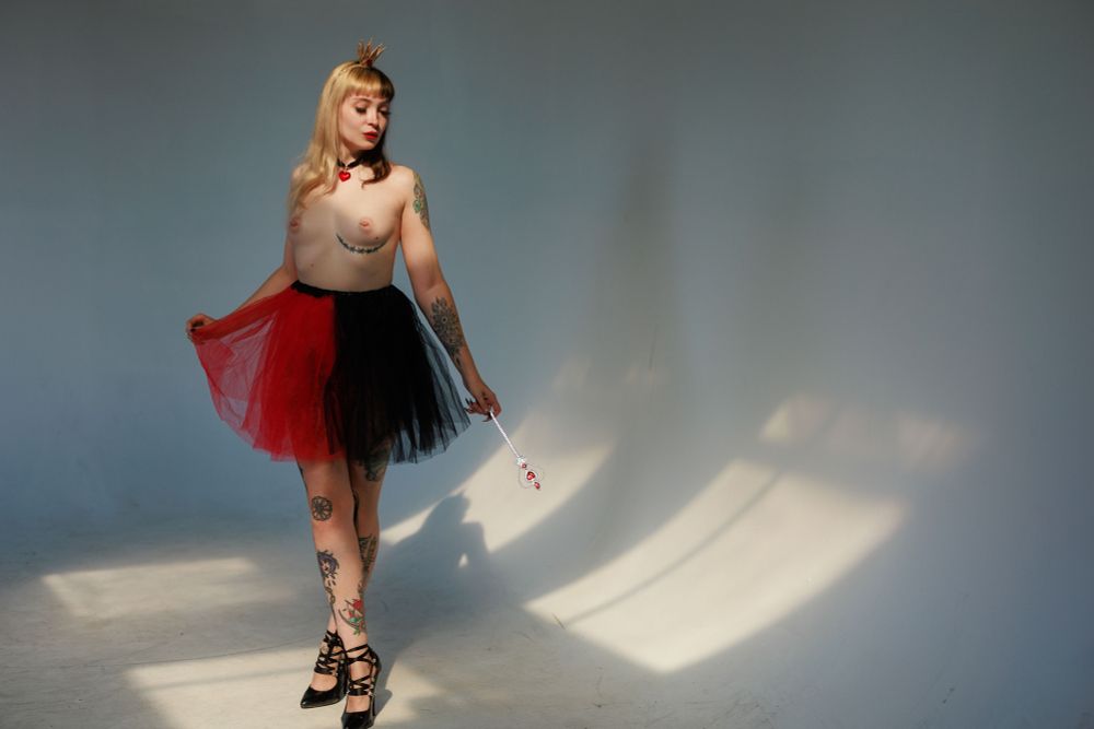 woman, standing, wearing tulle red-and-black skirt, crown, and carrying scepter (Queen of Hearts costume), topless, natural light from window