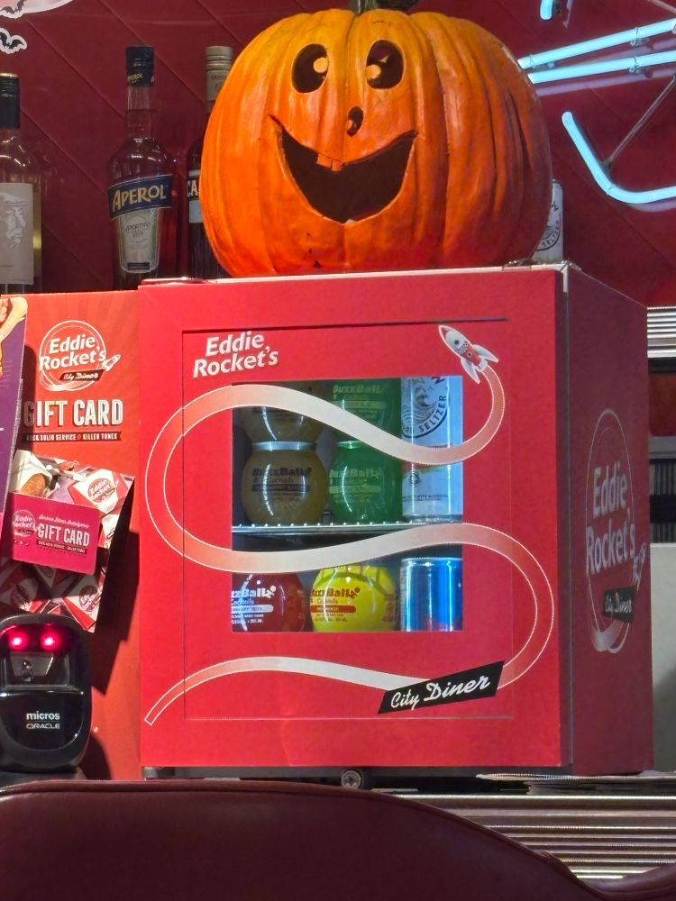 a small "Eddie Rocket's" branded fridge with buzzballz, white claw, and sugar free redbull in it, and a jack-o-lantern on top of it