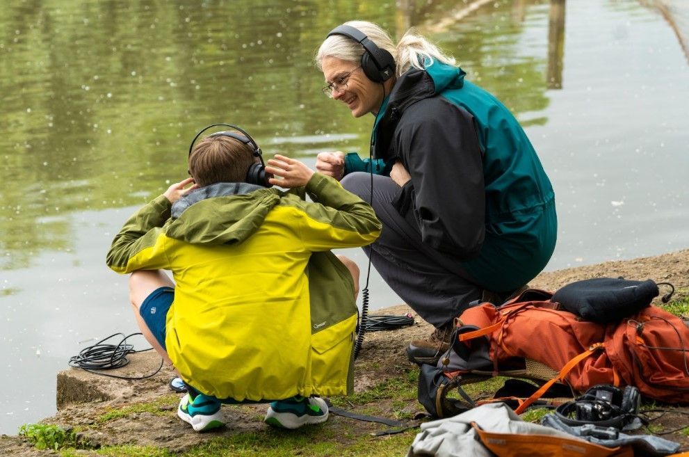 A woman and a boy wearing headphones crouch by a lake, listening and smiling. Backpacks and cables lie nearby on the ground.