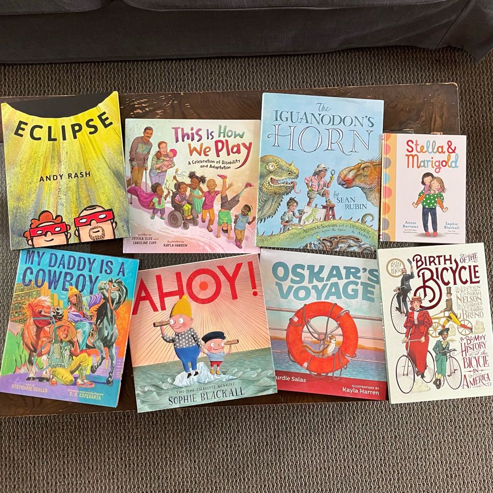 8 books on a table, including: ECLIPSE, THIS IS HOW WE PLAY, THE IGUANODON’S HORN, STELLA & MARIGOLD, MY DADDY IS A COWBOY, AHOY!, OSKAR’S VOYAGE, and BIRTH OF THE BICYCLE