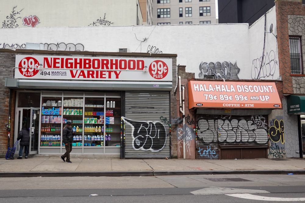 A man strolls by two neighboring Brooklyn storefronts - 99¢ Neighborhood Variety and Hala-Hala Discount. Each store has graffiti on their pulldown gates by SEO and CHEAK. NOTICE has painted on the store's rooftops.