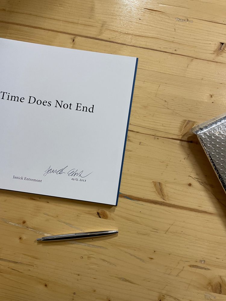 A signed copy of the book “If Time Does Not End” by Janick Entremont on a wooden table. The book’s title is centered on the page, with the author’s signature and date below. A silver pen lies nearby, next to bubble wrap packaging.