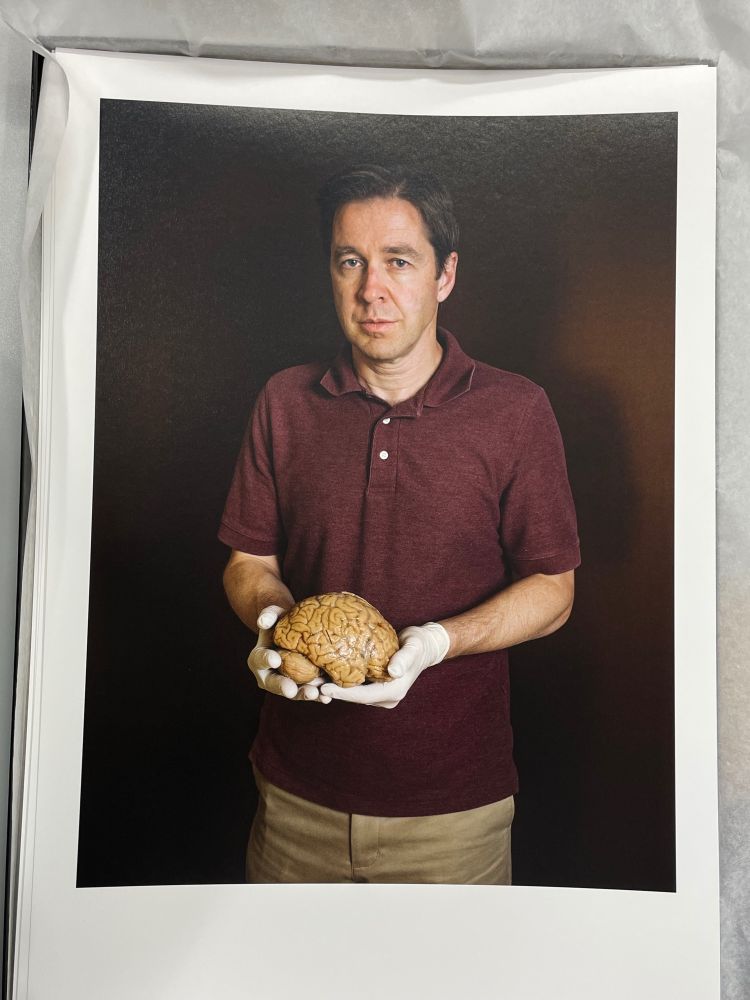 A close-up photo of a printed portrait featuring a man wearing a burgundy polo shirt and beige pants. He is holding a realistic human brain with both hands, wearing white gloves. The background is dark, creating a dramatic contrast that highlights the subject and the brain model. The man looks directly at the camera with a serious expression.