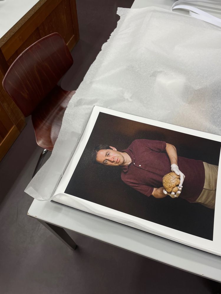 A photo of a printed portrait lying on a table, partially covered by protective tissue paper. The portrait shows a man wearing a burgundy polo shirt and holding a human brain while wearing white gloves. He stands against a dark background, looking directly at the camera. The table is in a room with wooden details, and a dark wooden chair is positioned next to it.