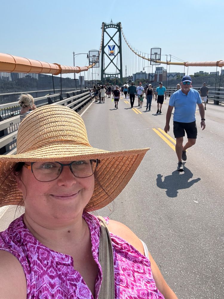 A person in a pink shirt and wide brimmed straw hat in front of a car suspension bridge with only people walking in the road