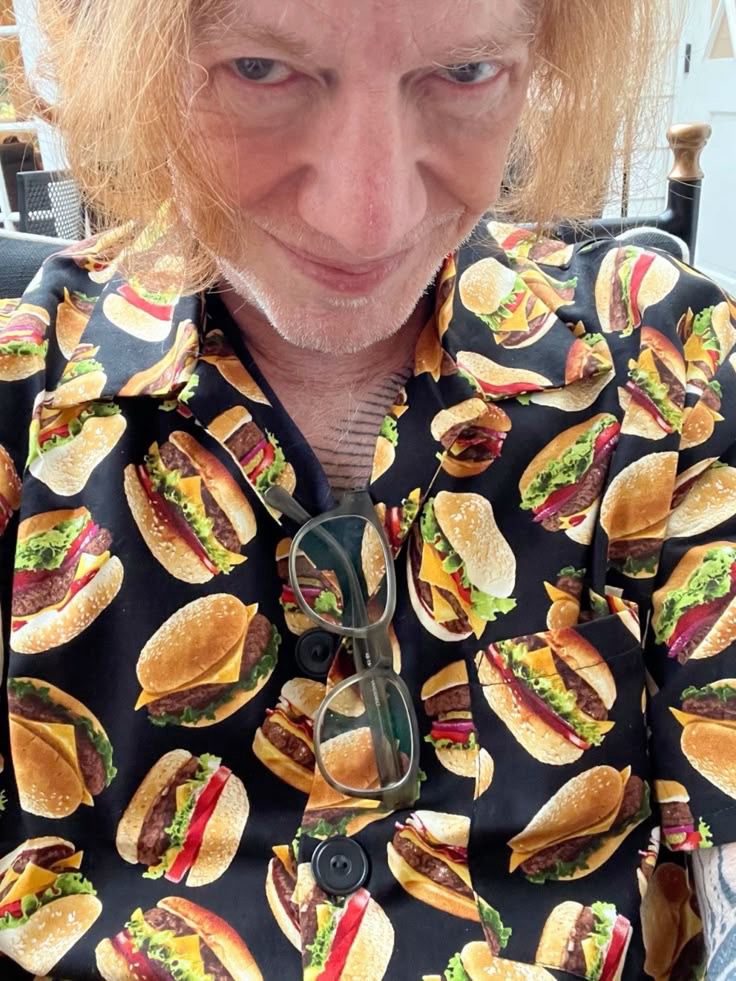 Danny Elfman smiling very silly and wearing a button up shirt with cheeseburgers all over it with glasses hanging in the middle of it.