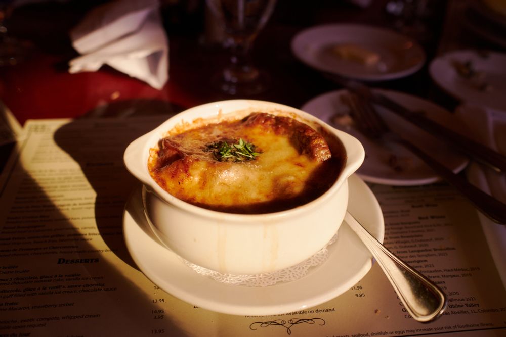 French onion soup set on a table. The bowl has gruyere cheese broiled on top with herbs centered on the cheese. A sliver of light illuminates the bowl