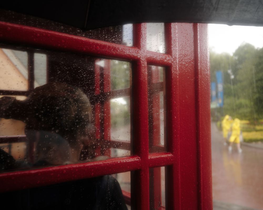 A woman taking refuge from the rain in a red telephone booth. People in yellow parkas are walking outside the booth at a distance.