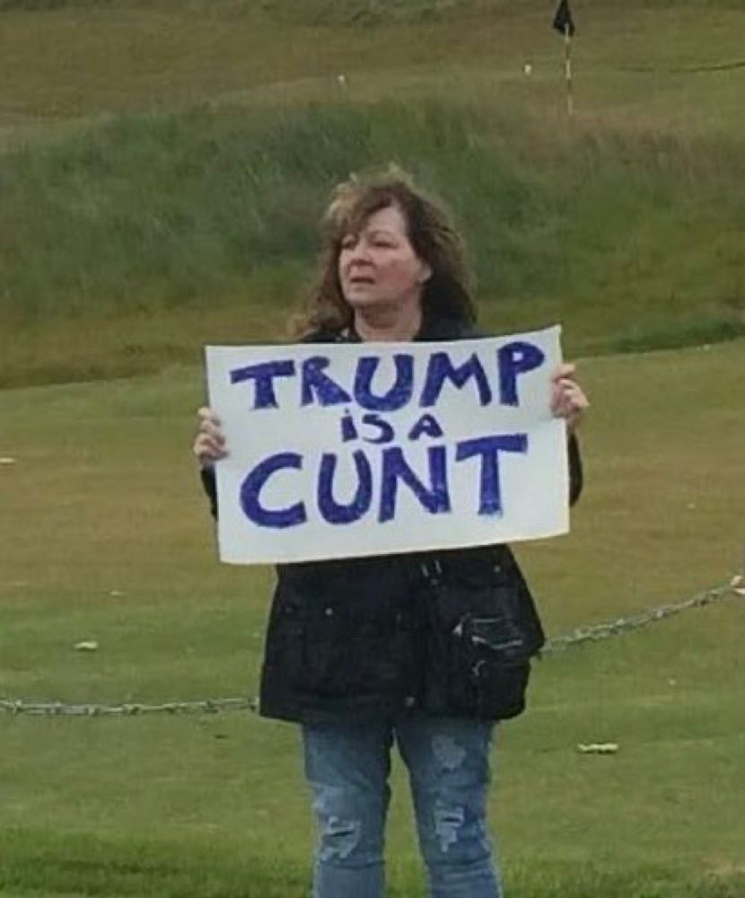 A picture of Janey Godley holding a placard describing an orange baboon on a Golf Course in Scotland