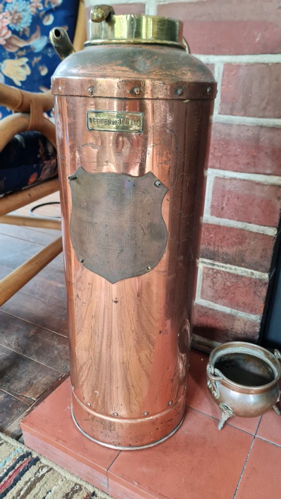Very old copper fire extinguisher with shield on the front (words not legible) a gold coloured tag saying Tested to 800lbs a handle and a hose connection