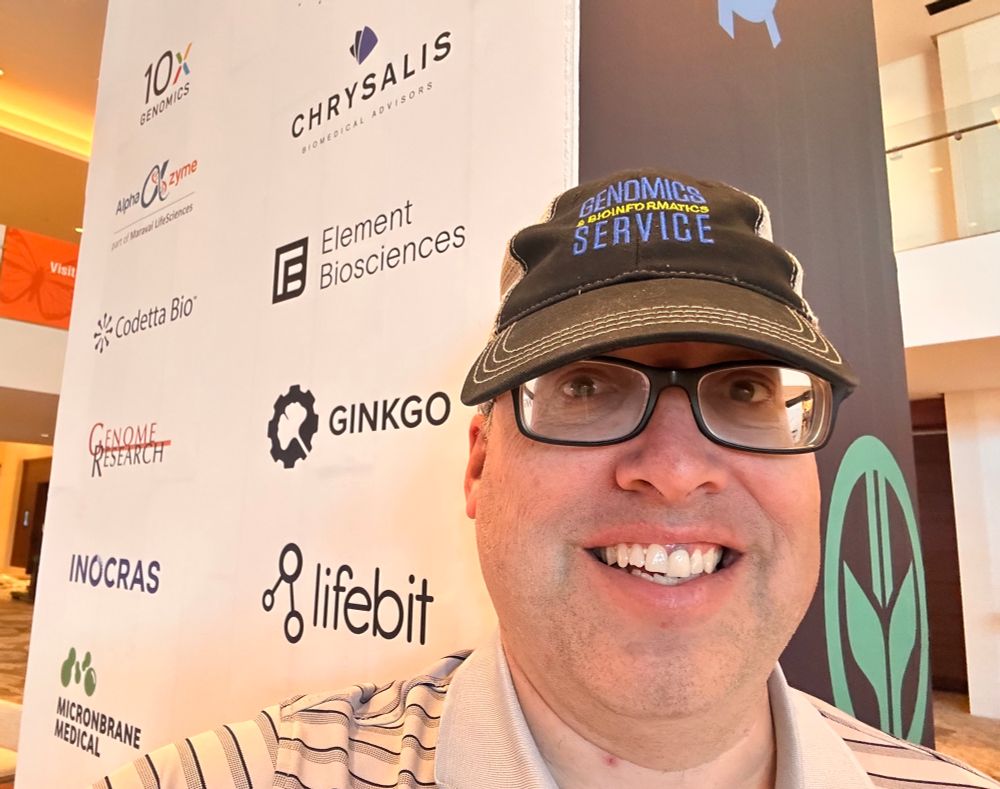 Photo of author wearing baseball cap lettered “Genomics & Bioinformatics Service” with AGBT sponsor list on pillar behind