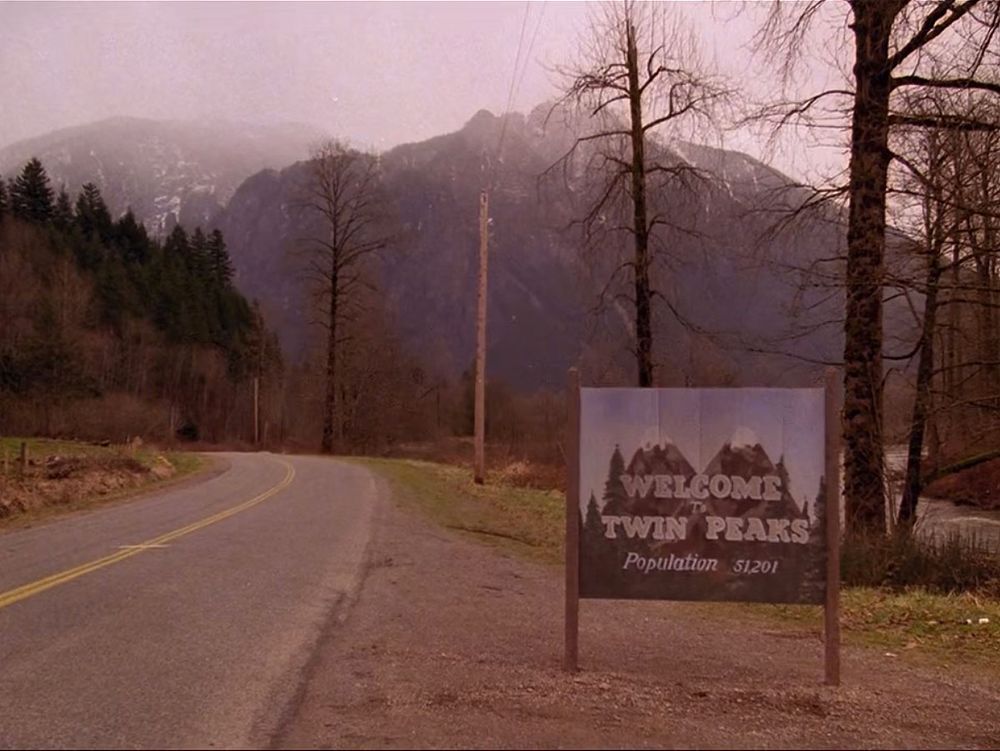 A still from the opening of the television series Twin Peaks.

A sign on the side of a two-lane road reads:

"Welcome To Twin Peaks
Population 51,201"

In the background are mountains and trees.