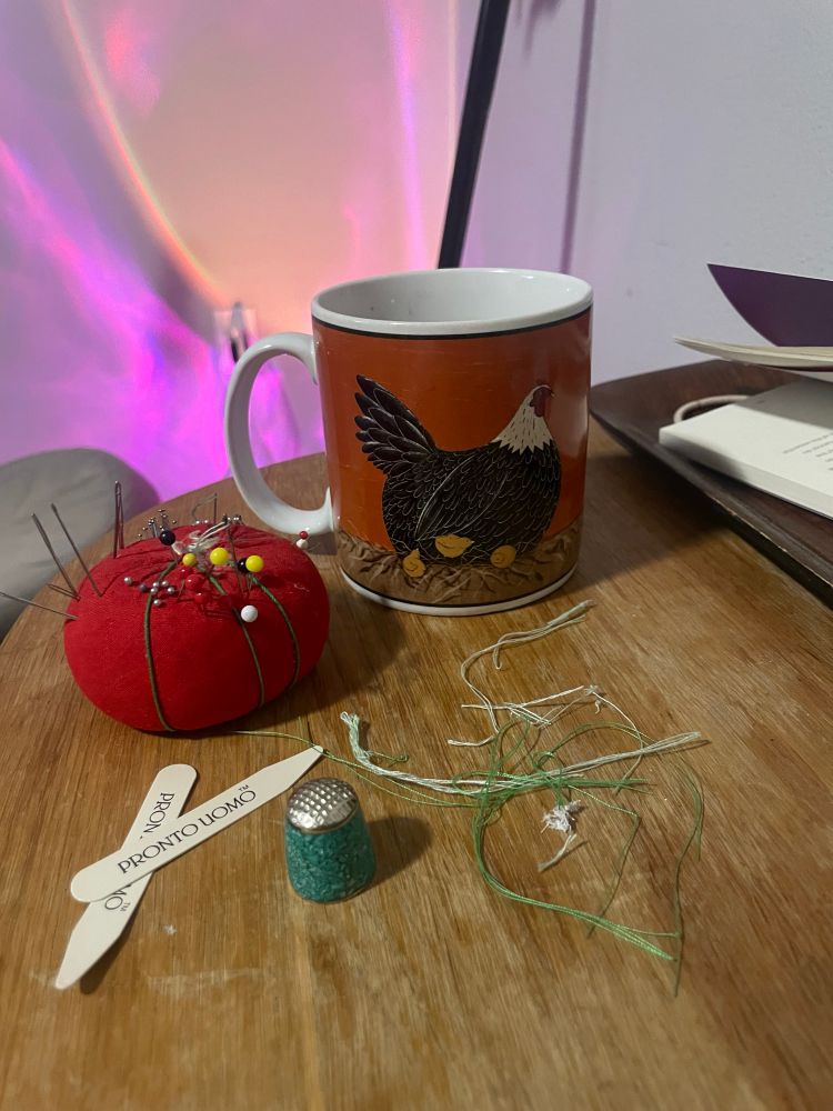 Sewing perephenalia and a chicken mug on a worn table with a pink light on the wall 