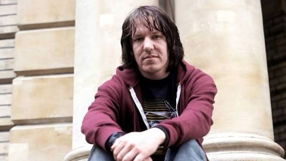 Elliott Smith sitting in front of some columns in a maroon hoodie