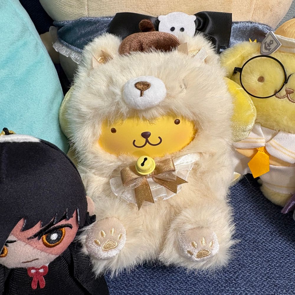 a picture of a pop mart pompompurin dressed as a fox bag charm. next to him is a black jack ballchain mascot and behind him is a pompompurin in a sailor suit and glasses plush.