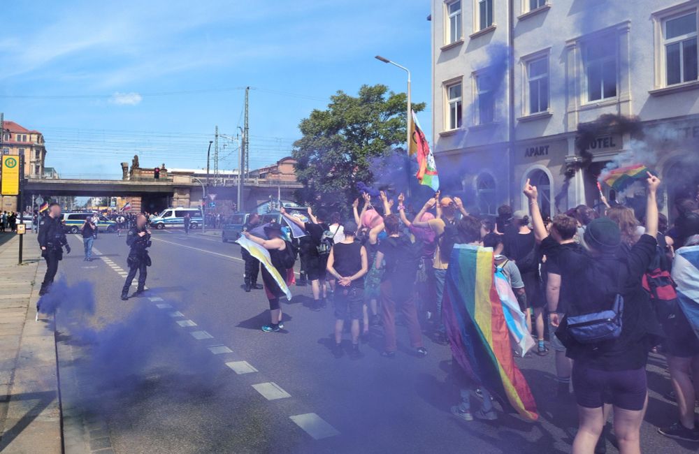 Eine Menge an Menschen mit Regenbogenflaggen steht auf der Straße und protestiert gegen einen vorbeilaufenden rechten Aufmarsch. Es ist lila und schwarzer Rauch in der Luft zu sehen.
