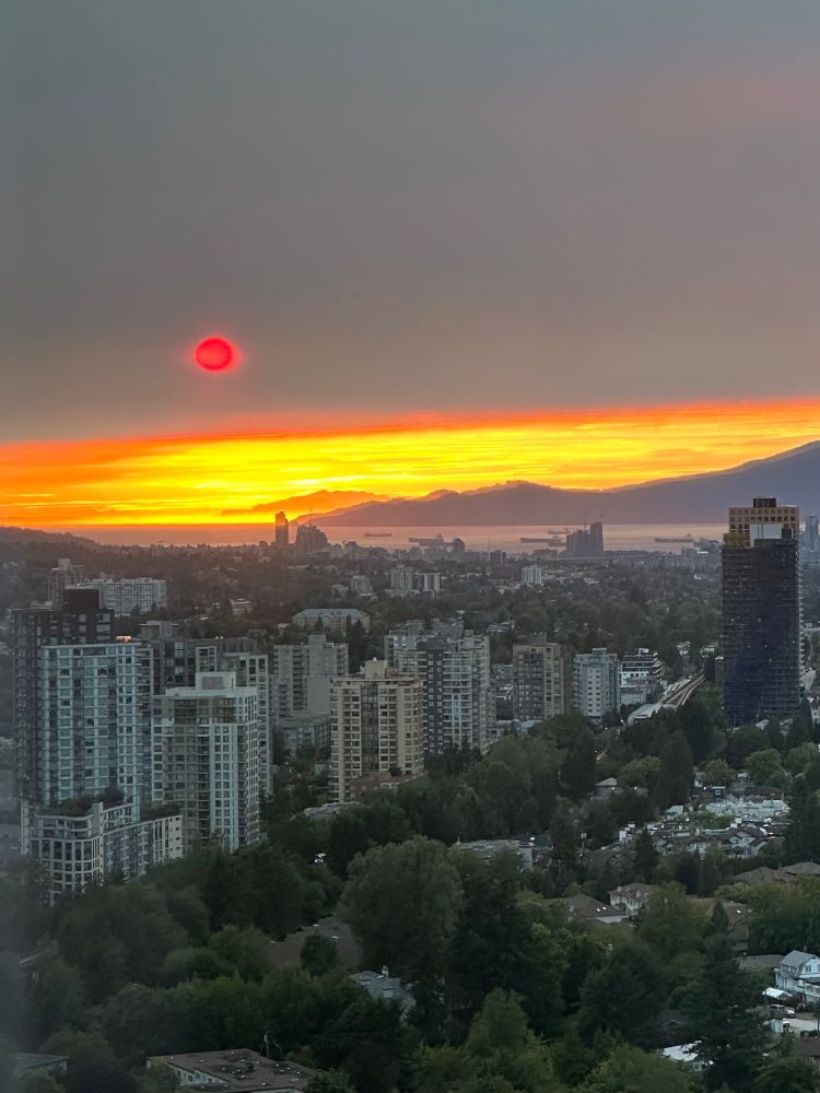 A landscape of Vancouver and English Bay. A bright red sun is setting against an incoming cloud of smoke. The water is a pale lavender and the sky at the horizon is bright yellow. 