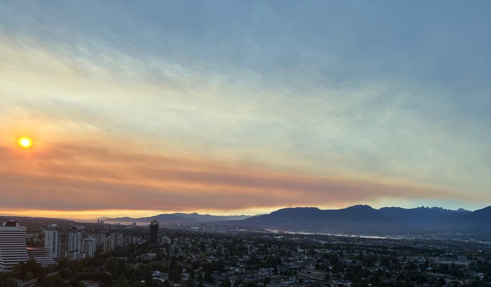 A panoramic view of the city of Vancouver and North Shore Mountains from 7:45pm Aug 12. The sky is blue but obscured by an incoming cloud of smoke. The sun is obscured, giving an eerie sunset glow an hour before sunset. 