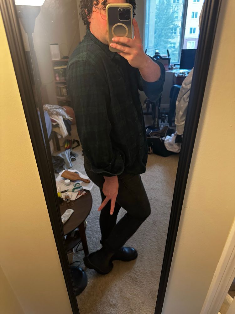 me taking a mirror selfie where i am cosplaying as kaite, i am taking the photo from the side wearing black combat boots, black skinny jeans, and a dark blue and green plaid flannel. my left hand is holding my phone in front of my face and my right hand is down by my ass in a peace sign 
