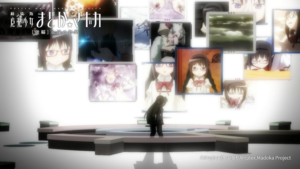 Homura is hugging Madoka in her room full of floating TVs showing her past self, with weird, flat uncomfortable furniture all around them. It's supposedly a couch but it looks more like multi colored benches, with nothing on them.
