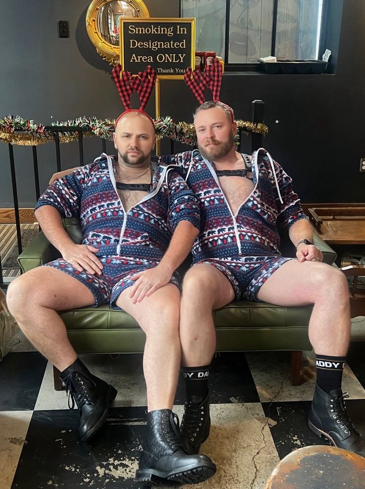 Two men in onesies with reindeer antlers pose 