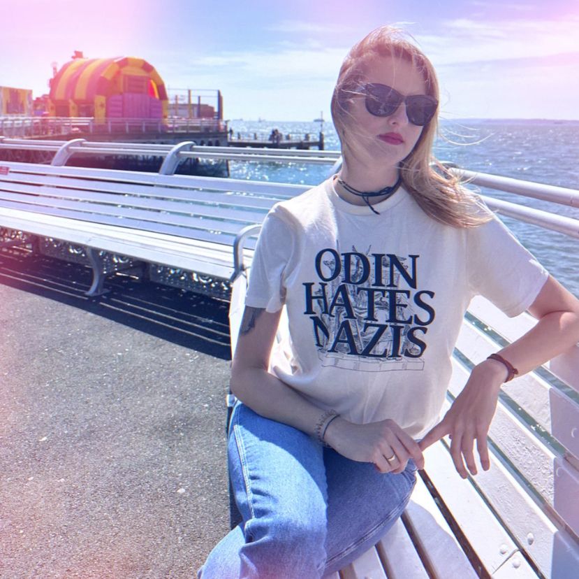 Woman with sunglasses on, sits on a bench with a pier behind her, wearing an Odin Hates Nazis t-shirt