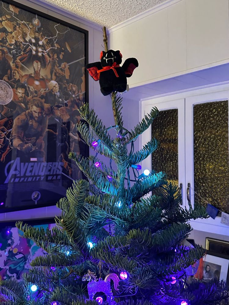 A small stuffed bat cat, who is black with red inside of his wings and inside of his ears and a red bow around his neck is at the top of a Christmas tree that has purple and white lights. There’s enough avengers infinity war poster behind the Christmas tree on the wall.
