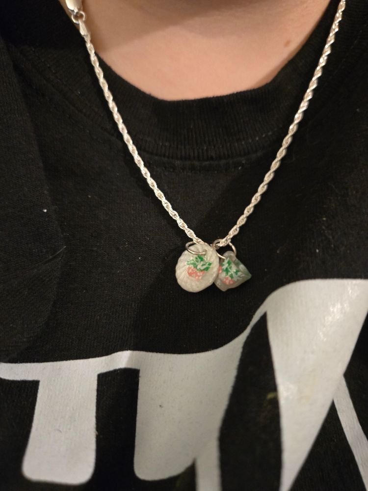 A photo of megan wearing the silver chain necklace. The two charms are of a white tea cup and white tea saucer that have painted strawberry and vine motifs on them. The cup and saucer are two separate charms hanging next to each other 
